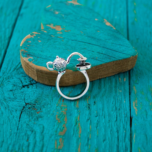 Silver teapot-shaped ring on a wooden surface with teal paint.